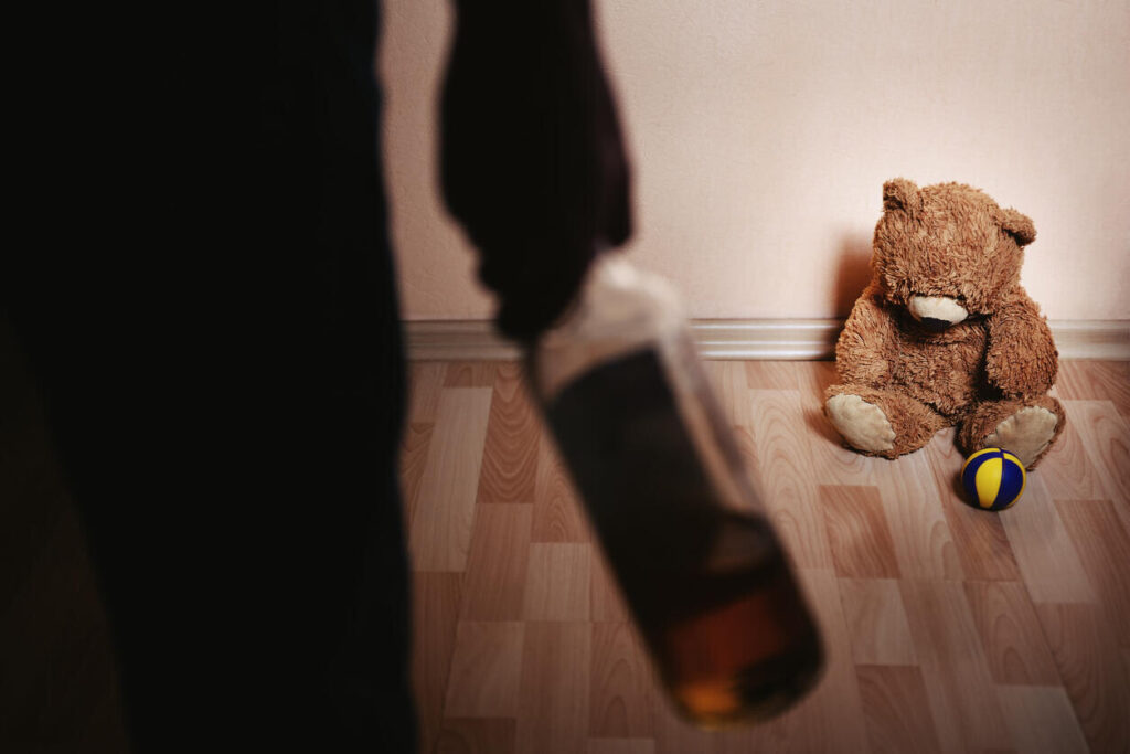 A shadowy figure holding a bottle stands near a teddy bear and a ball on the floor, symbolizing the impact of domestic assault allegations and the need for defense in Michigan.